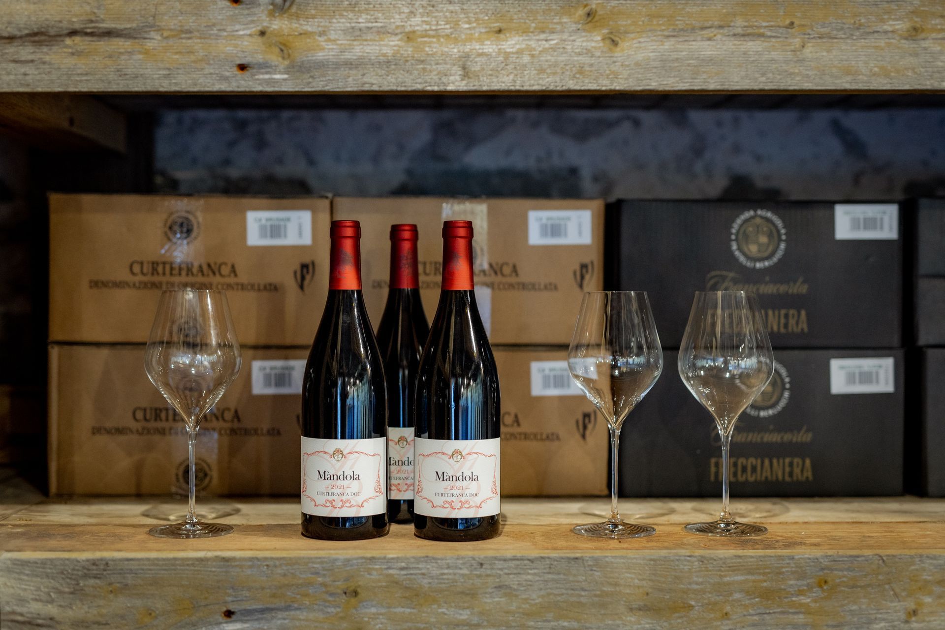 Wine bottles and glasses on a wooden shelf, with boxes of wine in the background.