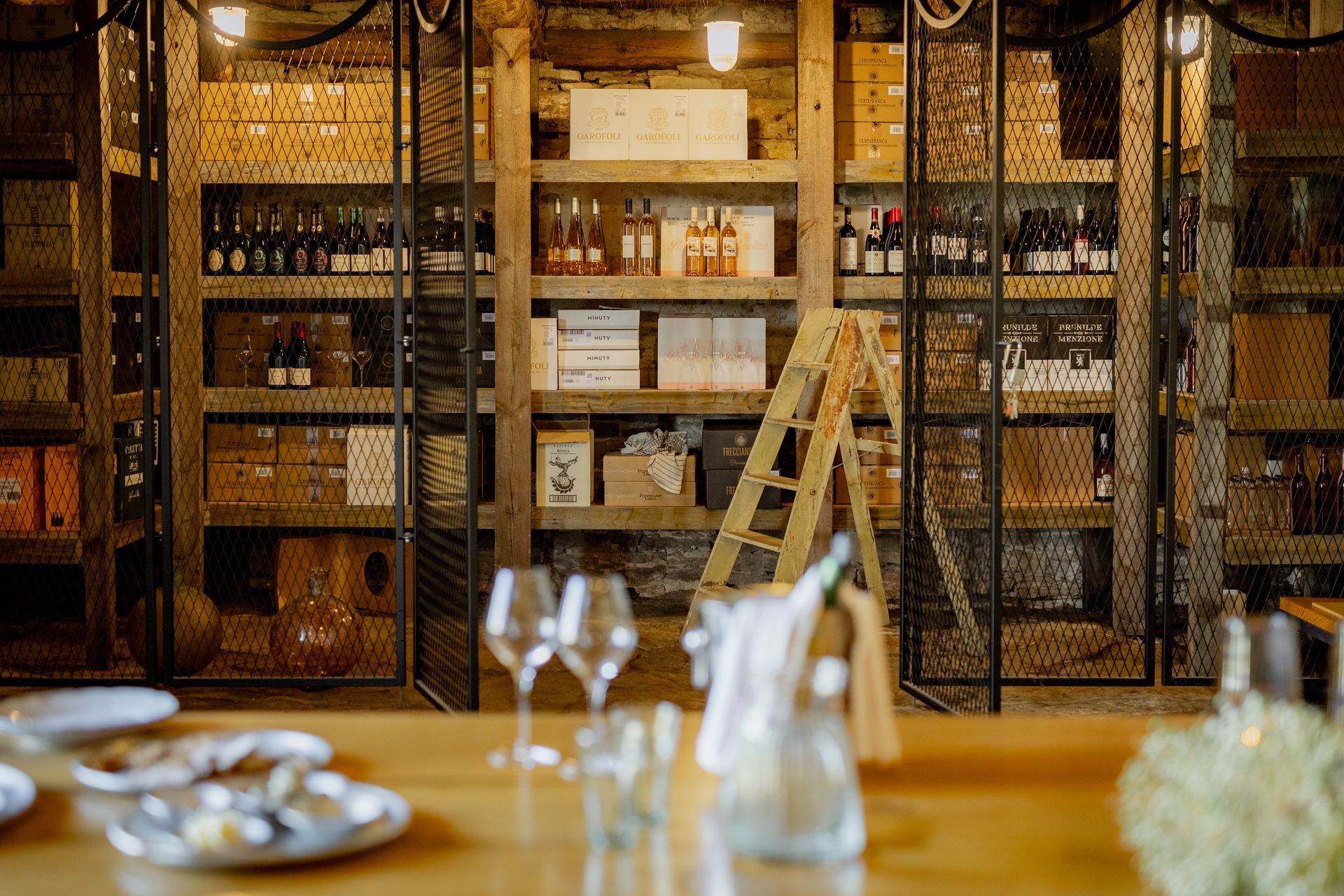 Wine cellar with wooden shelves, bottles, boxes, and a ladder; table set in foreground.