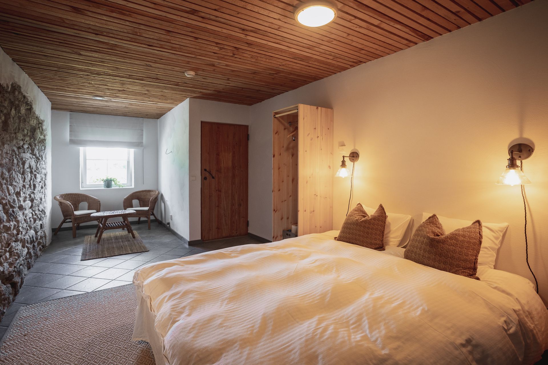 Bedroom with stone wall, wooden ceiling, bed, chairs, and lamps.