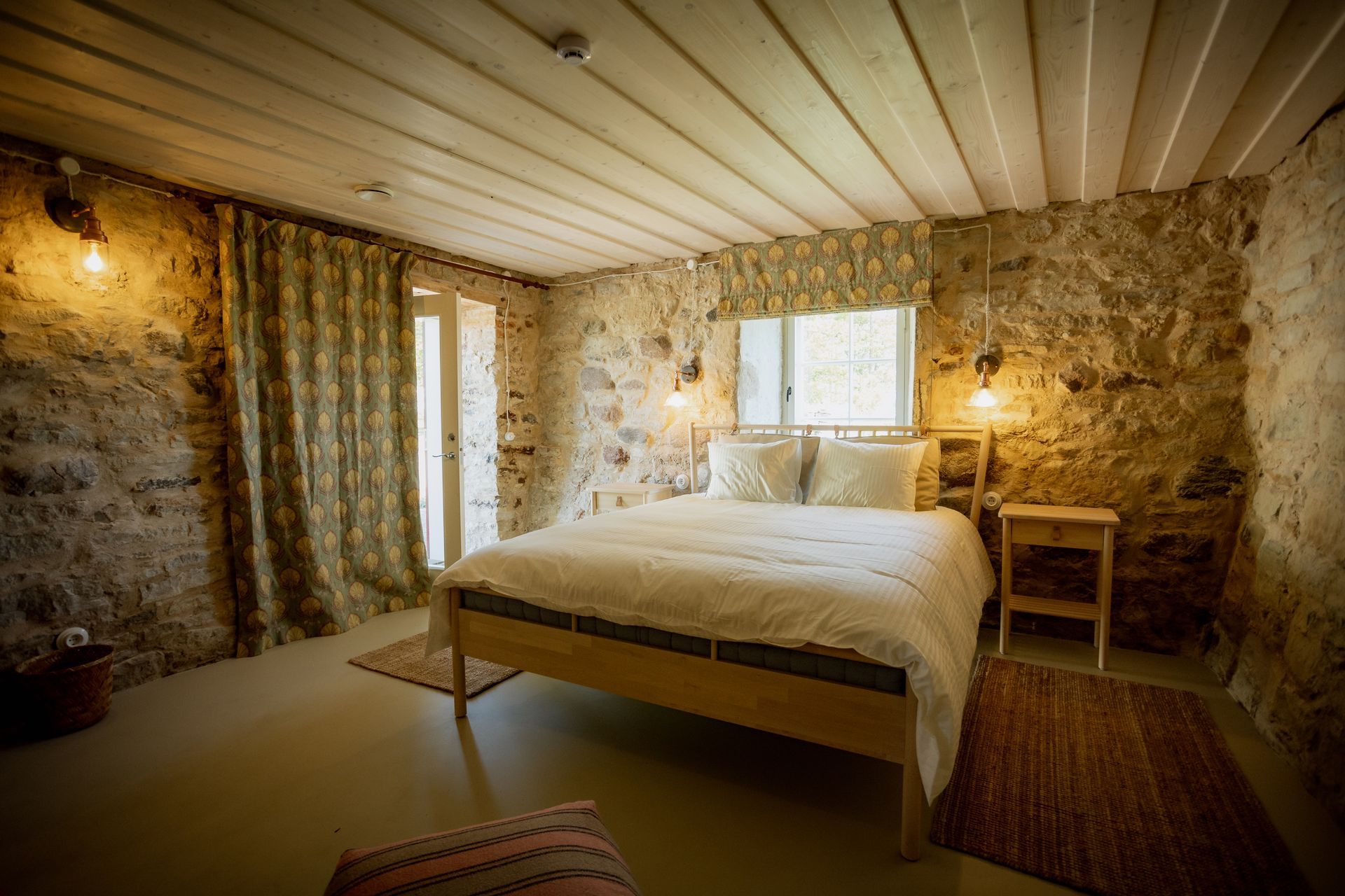 Bedroom with stone walls, wooden ceiling, bed, window, and two small bedside tables with lamps.