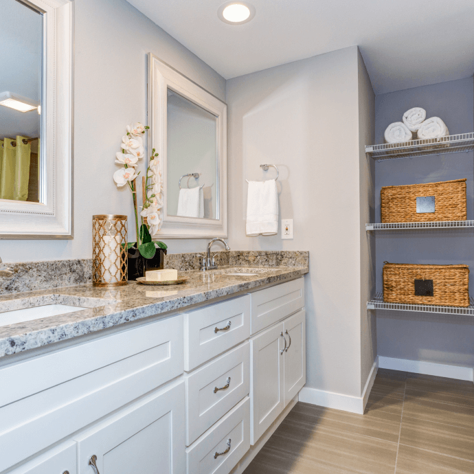 Bright bathroom vanity with marble counter, white cabinets, mirror, and wicker baskets on open shelving.