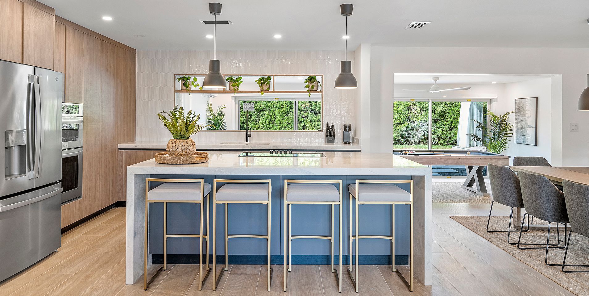 Bright modern kitchen with blue island stools, stainless fridge, and open dining area by large windows