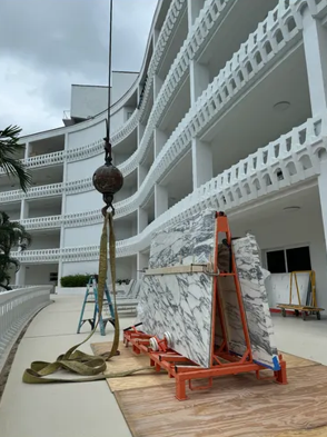 Construction site beside a white curved building, with marble slabs on orange carts and a crane hook overhead