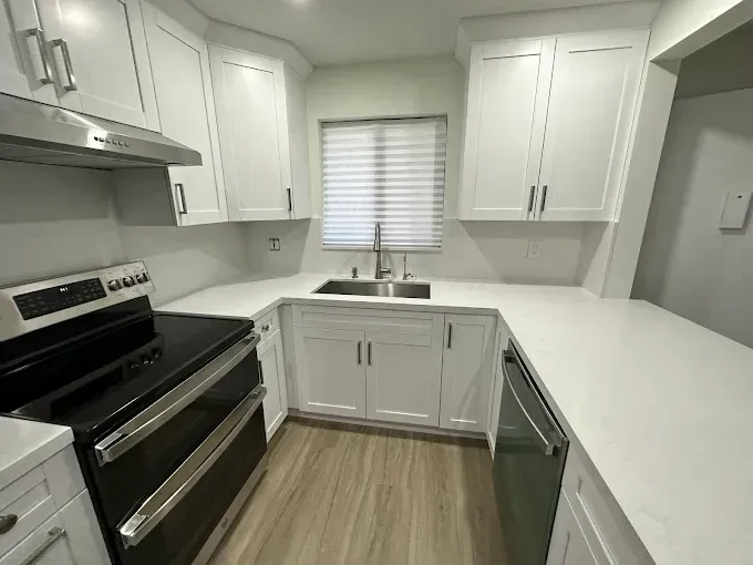 Small white kitchen with black appliances, gray wood floor, and a window over the sink.