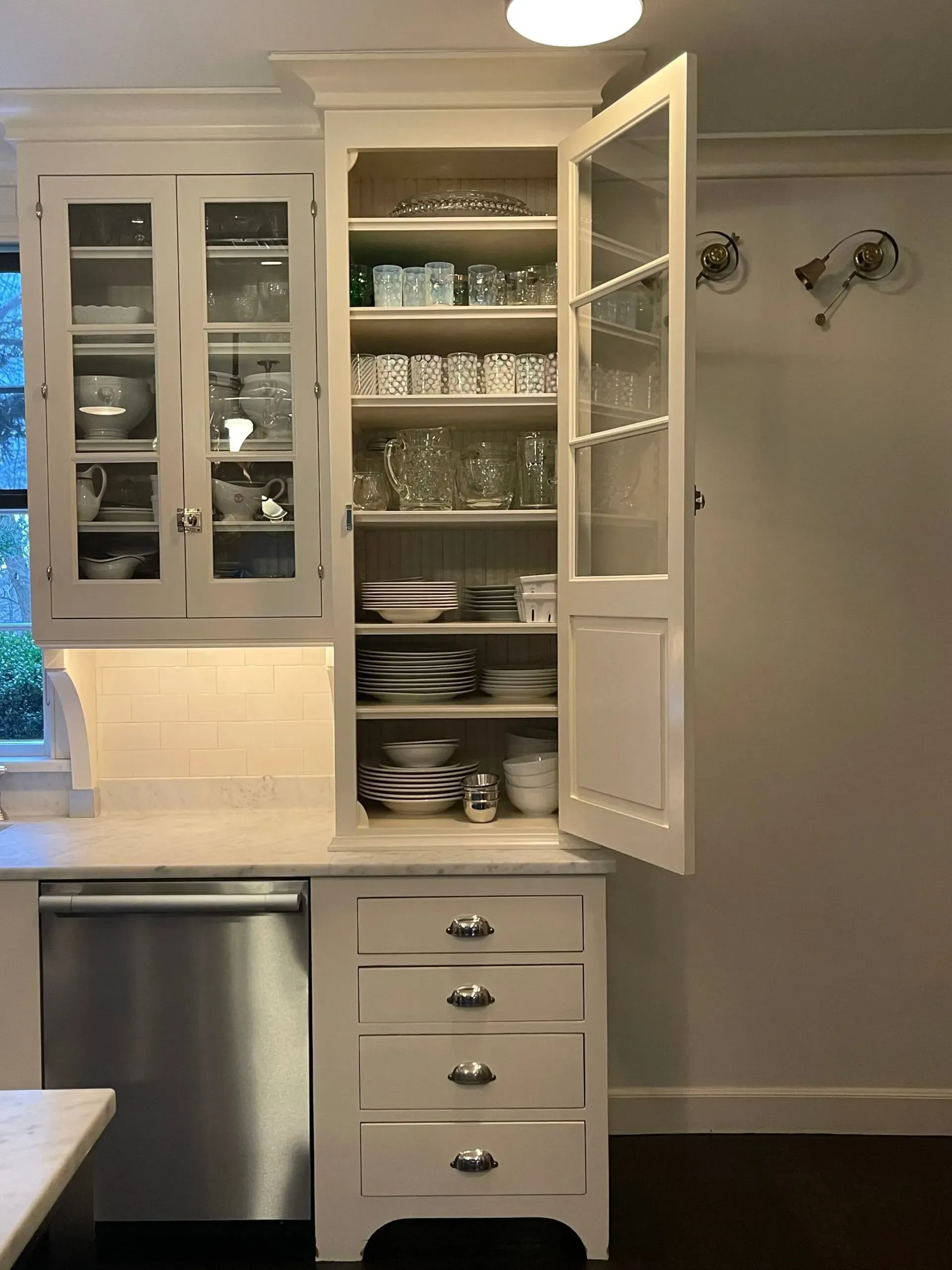 White kitchen cabinet with open glass doors showing dishes and glassware, next to a dishwasher.