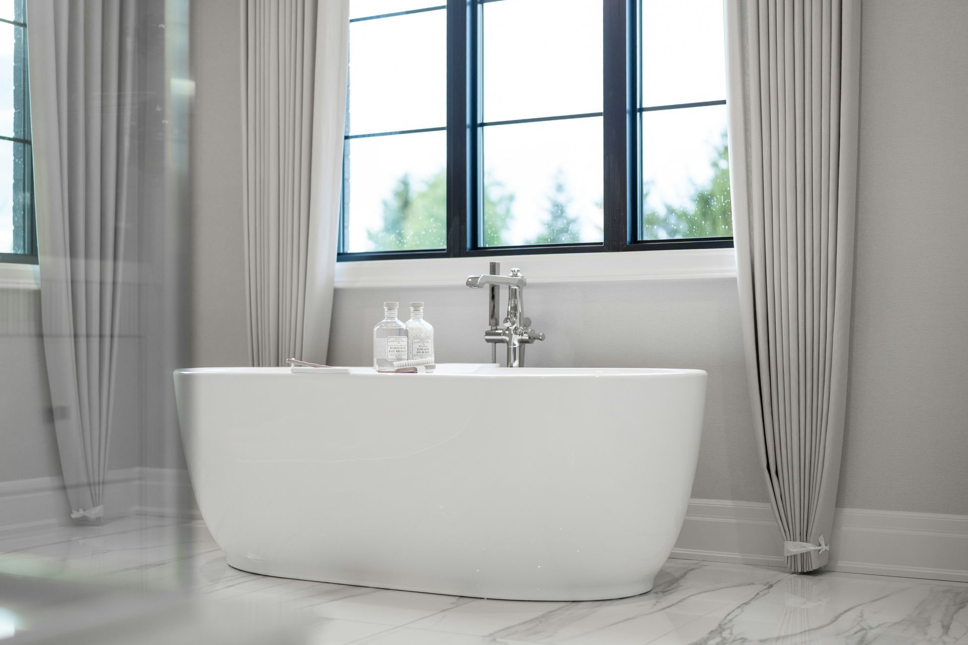 Modern bathroom with a freestanding white tub beneath large windows and gray curtains