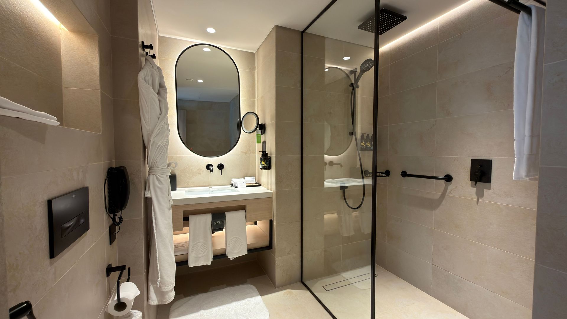 Modern bathroom with vanity, oval mirror, glass shower, and beige stone walls