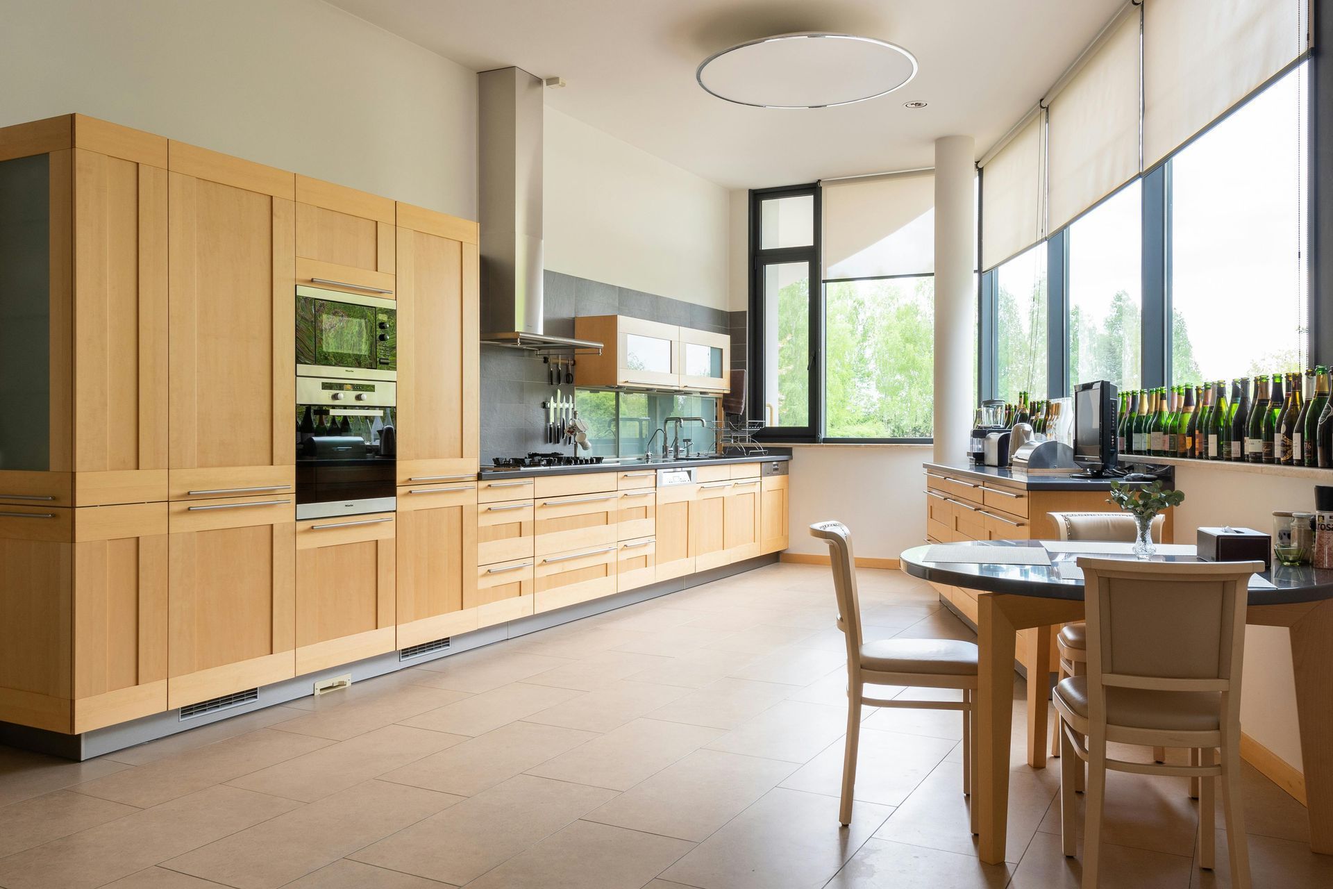Modern kitchen and dining area with light wood cabinets, large windows, and a table by the wall