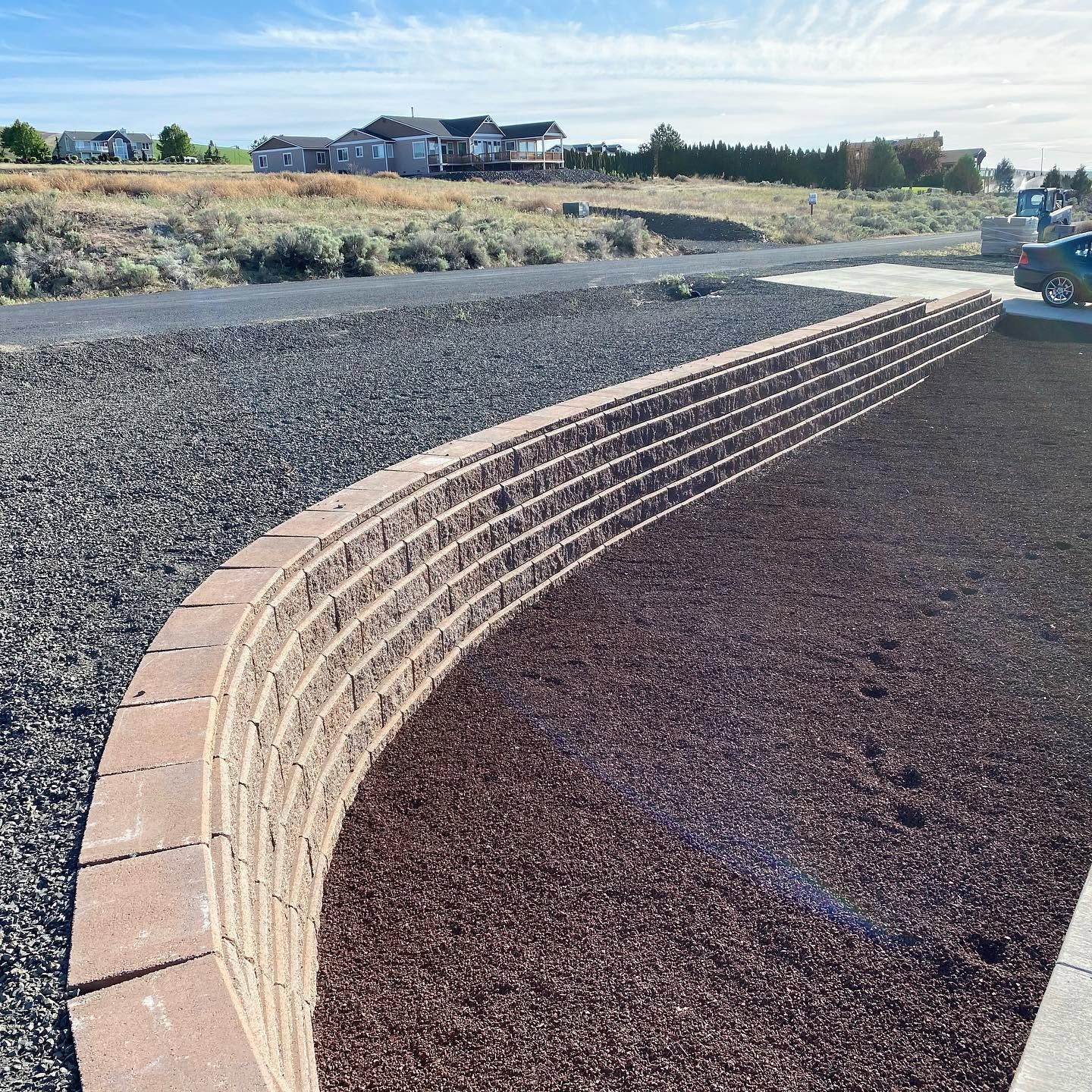 Curved brick retaining wall beside a gravel lot with a road and houses in the distance.
