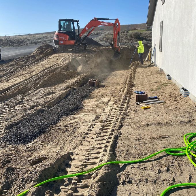Excavator digging beside a building on a dirt construction site, with a worker nearby and hoses in the foreground
