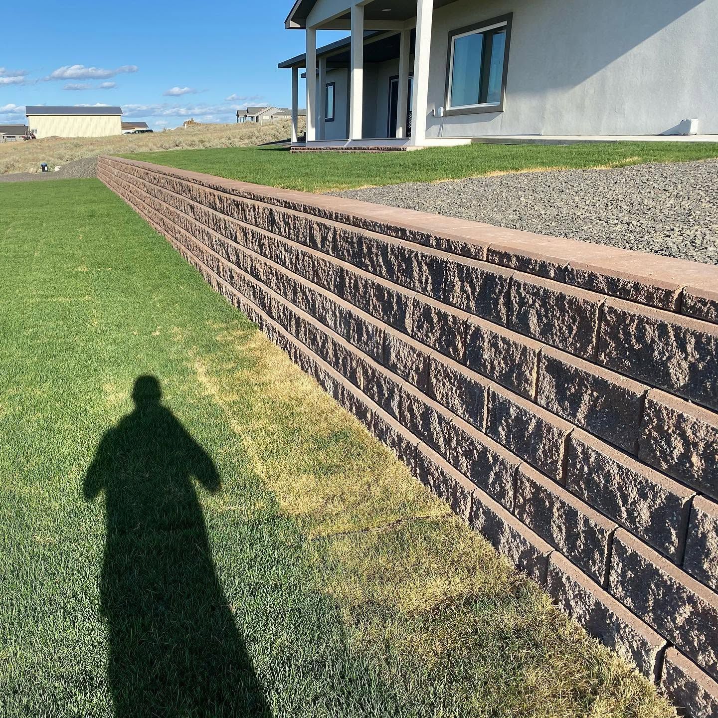 House with stone retaining wall beside a green lawn under a clear sky, with a person’s shadow in the foreground