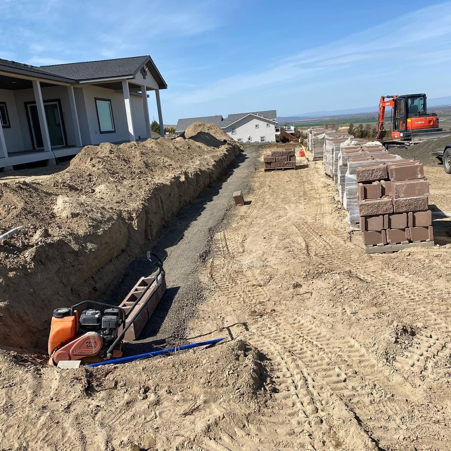 House under construction with dirt trenches, stacked bricks, and an excavator in a suburban lot.