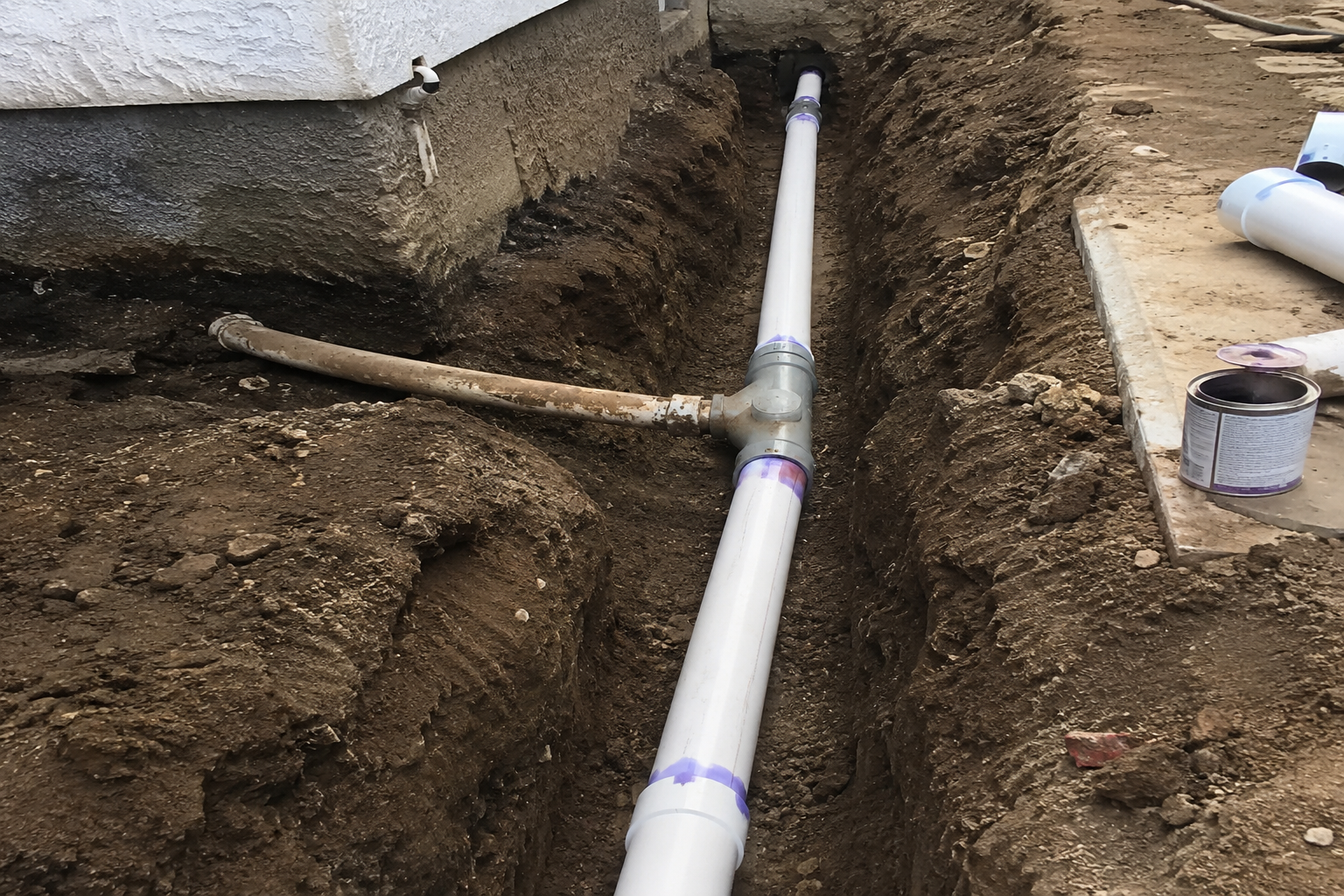 Trench with white PVC pipe laid in dirt beside a house foundation and loose soil.