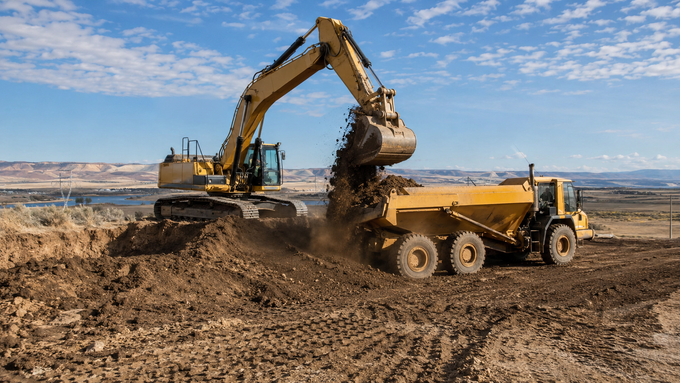 Yellow skid steer pushing a mound of dirt on a bare lot with trees in the background