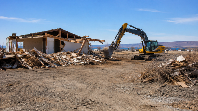 Excavator demolishing a house, with rubble and debris collapsing beside neighboring buildings.