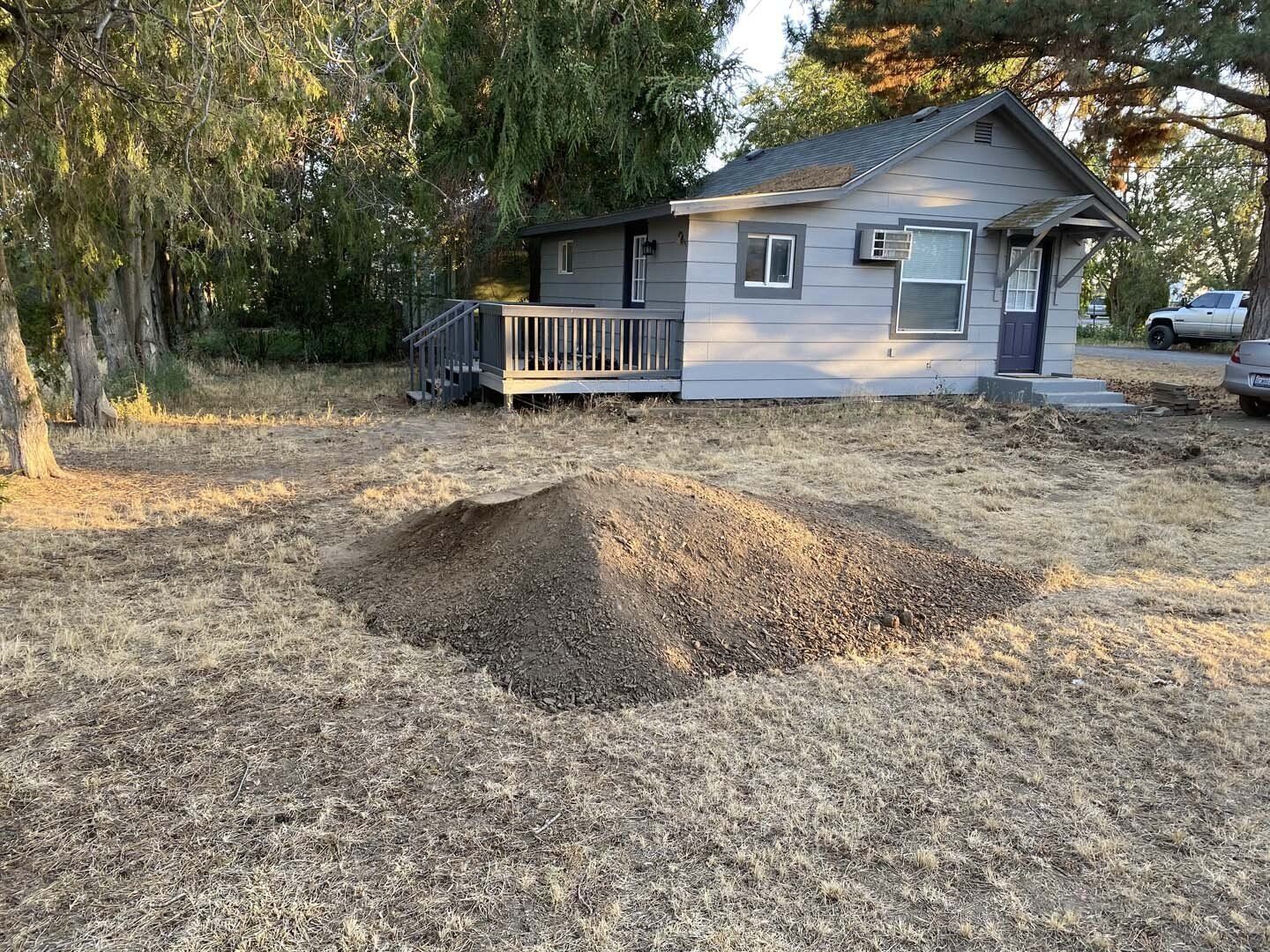 A large dirt mound in a yard in front of a small gray house with trees around it.