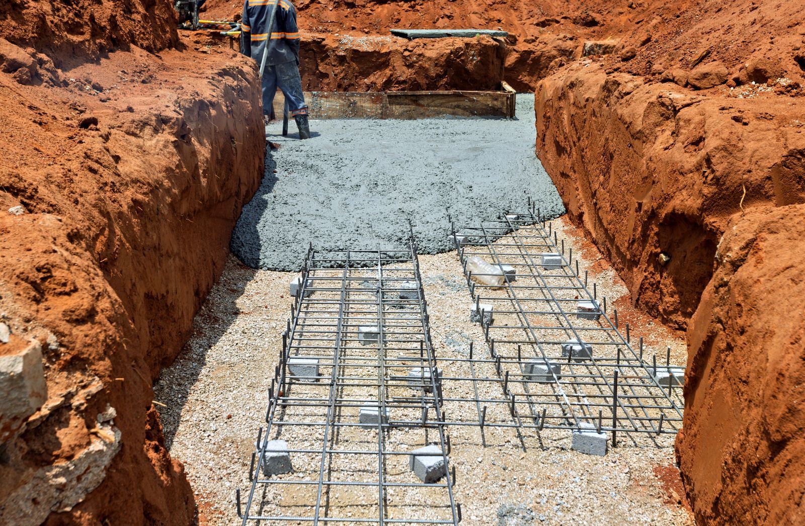 Construction trench with rebar grid and gravel foundation, worker standing at the far end.