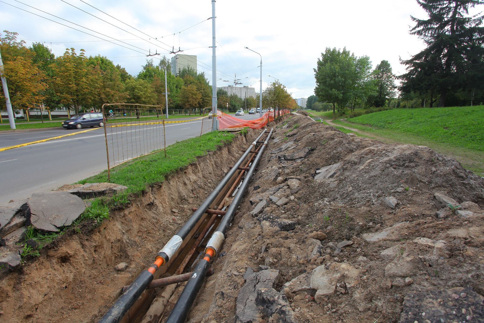 Utility pipes laid in a roadside trench beside a highway, with orange safety fencing and dirt excavation.
