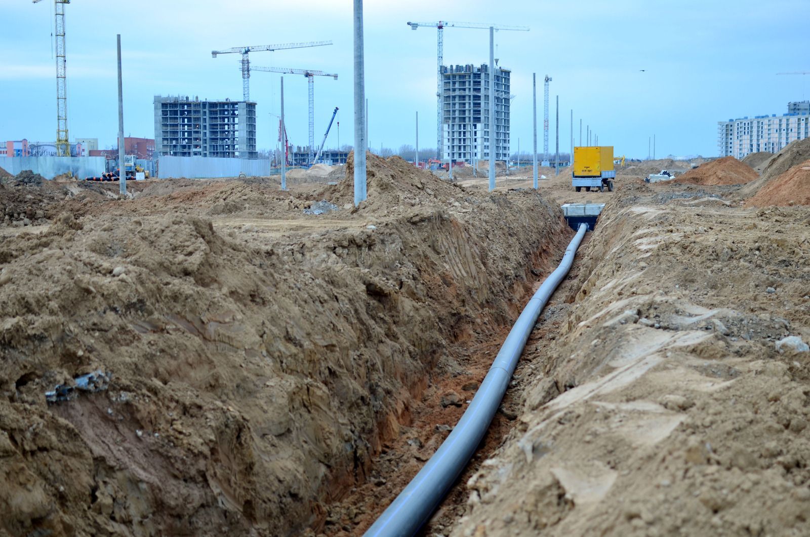 Construction site with trench and large black pipeline leading toward distant buildings and cranes