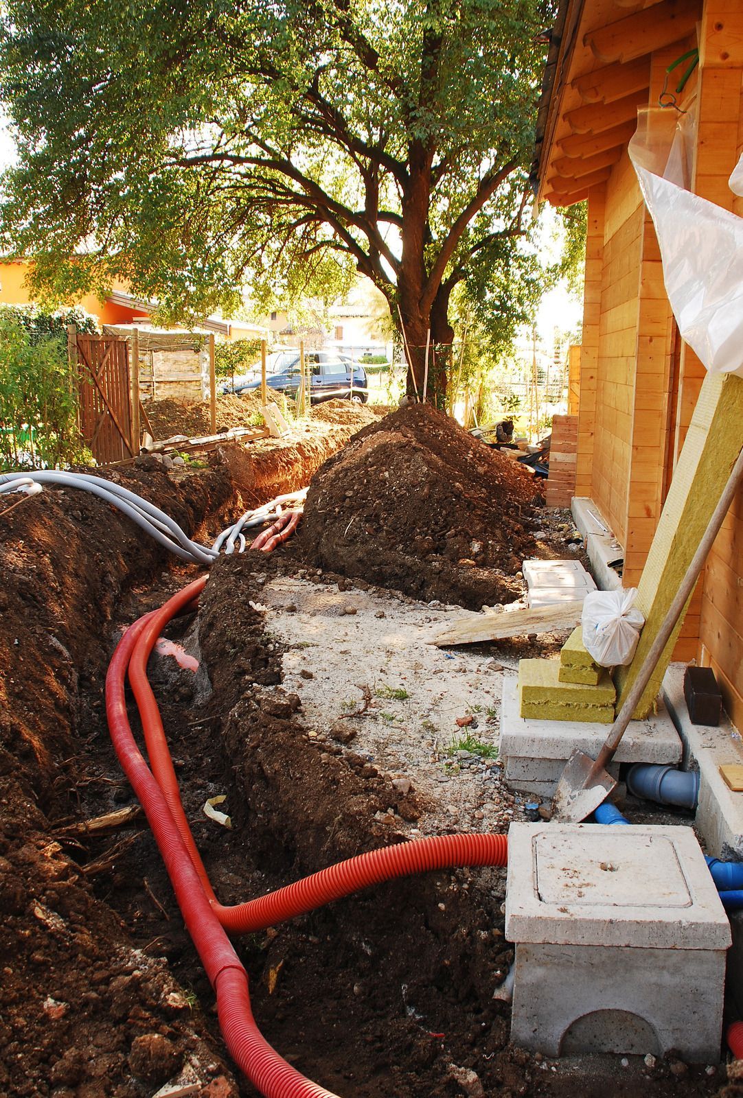 Trenches and red pipes beside a house foundation, with dirt piles and a tree overhead