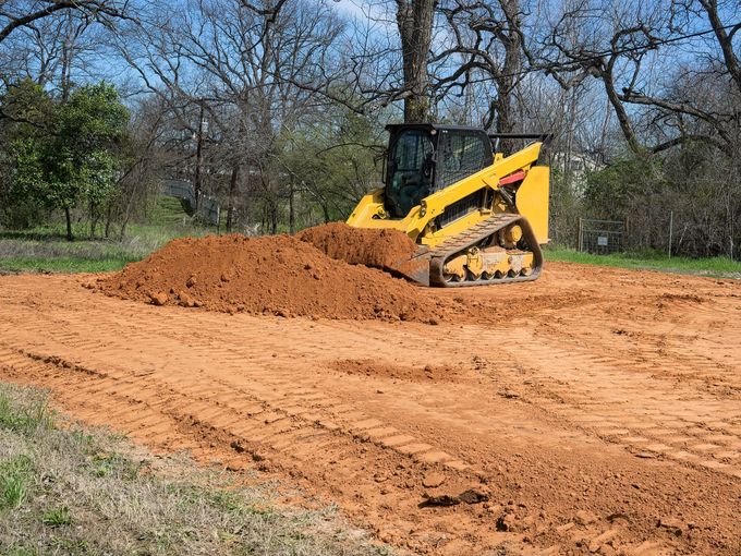 Yellow skid-steer loader moving dirt on a cleared lot with trees in the background