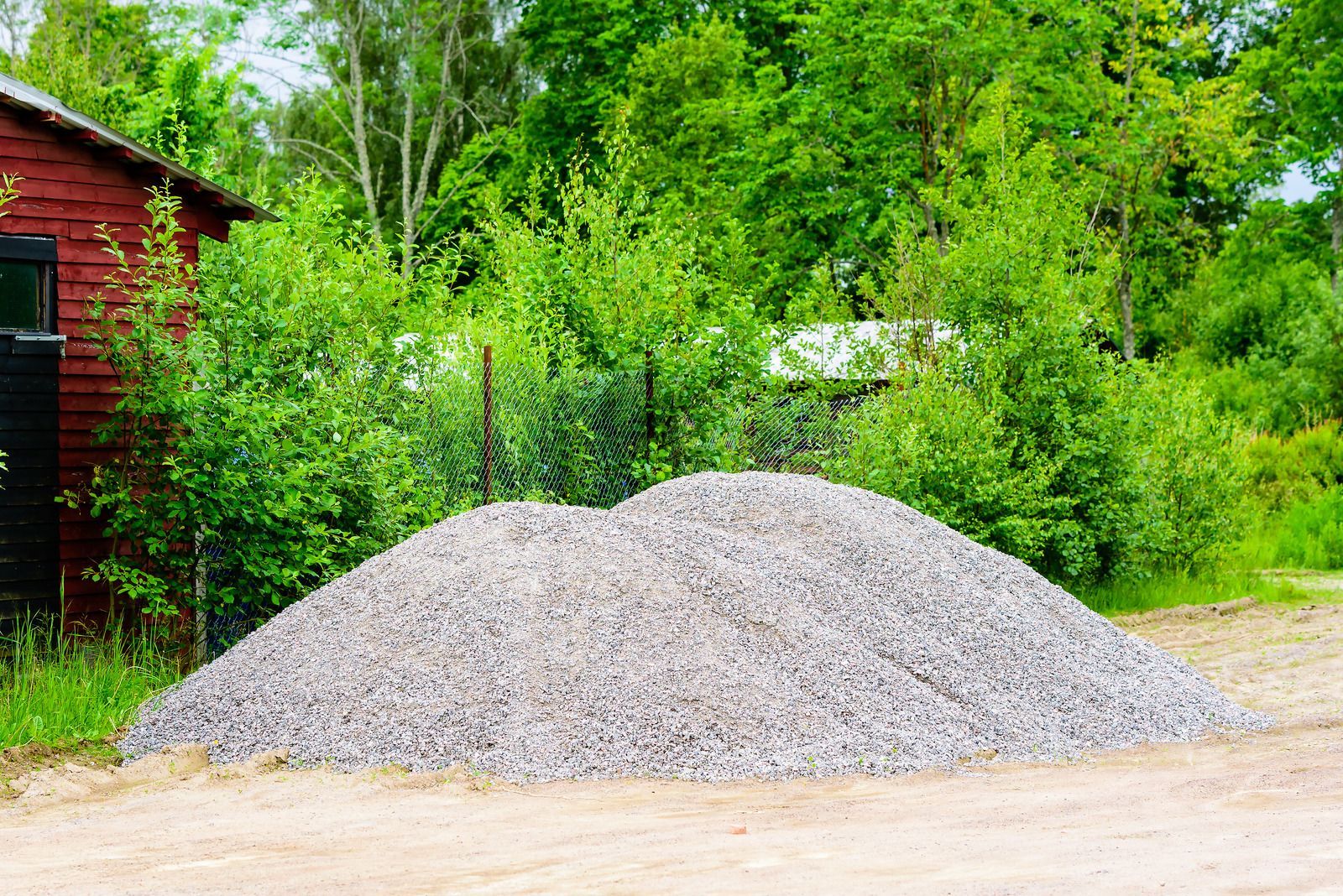 Large gravel pile in front of a red building, with green trees and brush in the background