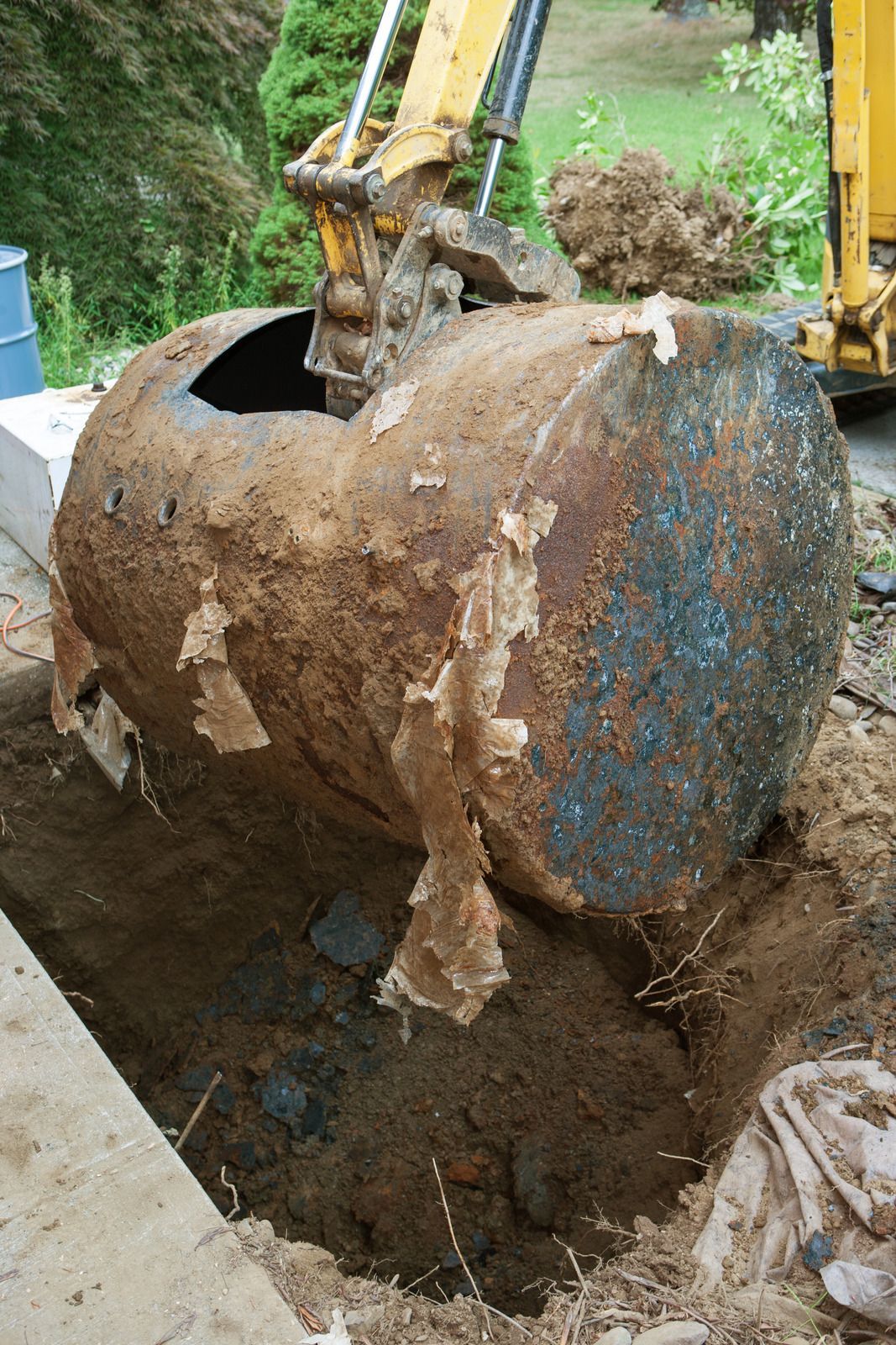 Excavator lifting a rusted cylindrical metal tank from muddy ground near a construction site.