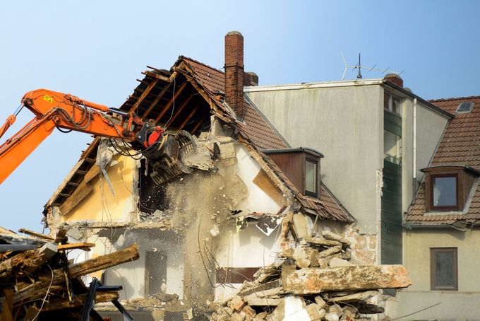 Excavator demolishing a house, with debris falling beside neighboring buildings under a clear sky