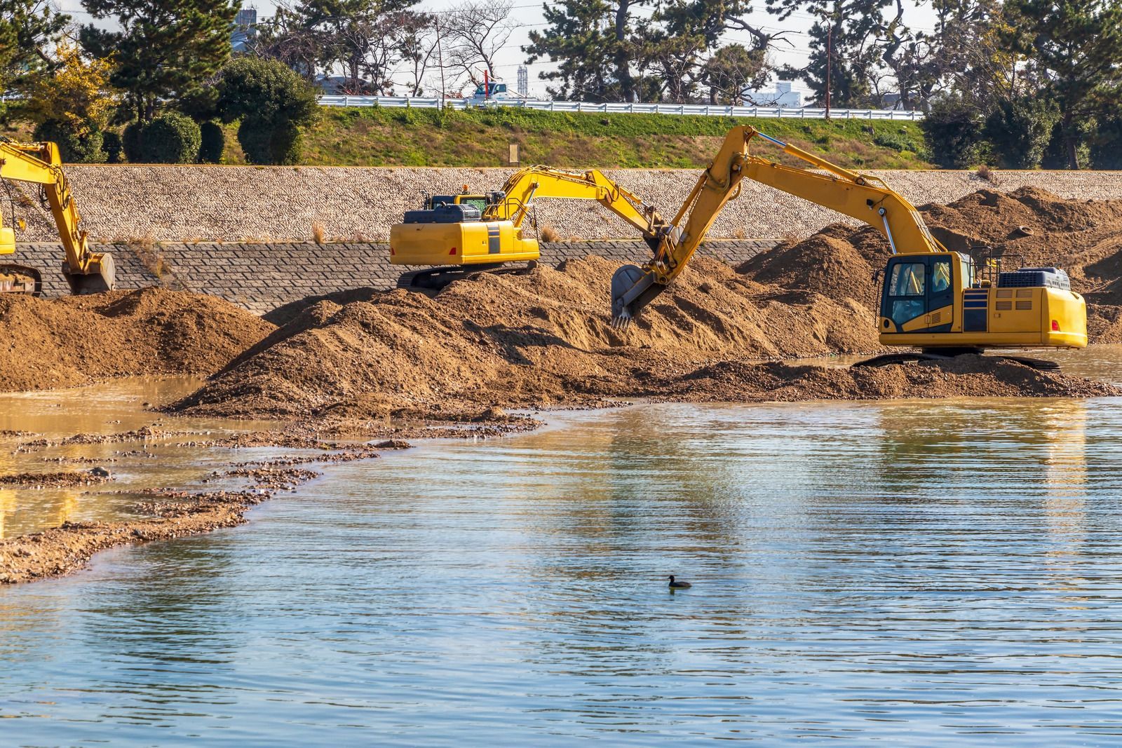 Two excavators digging dirt near a riverbank, with water in the foreground and trees behind.