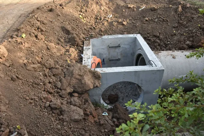 Concrete drainage culvert under construction in a dirt excavation, with a worker in orange gear nearby