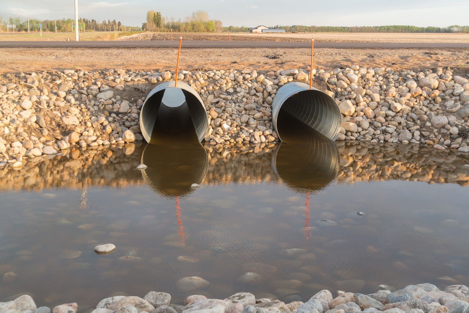 Two large drainage pipes empty into a rocky retention pond with still water at sunset.