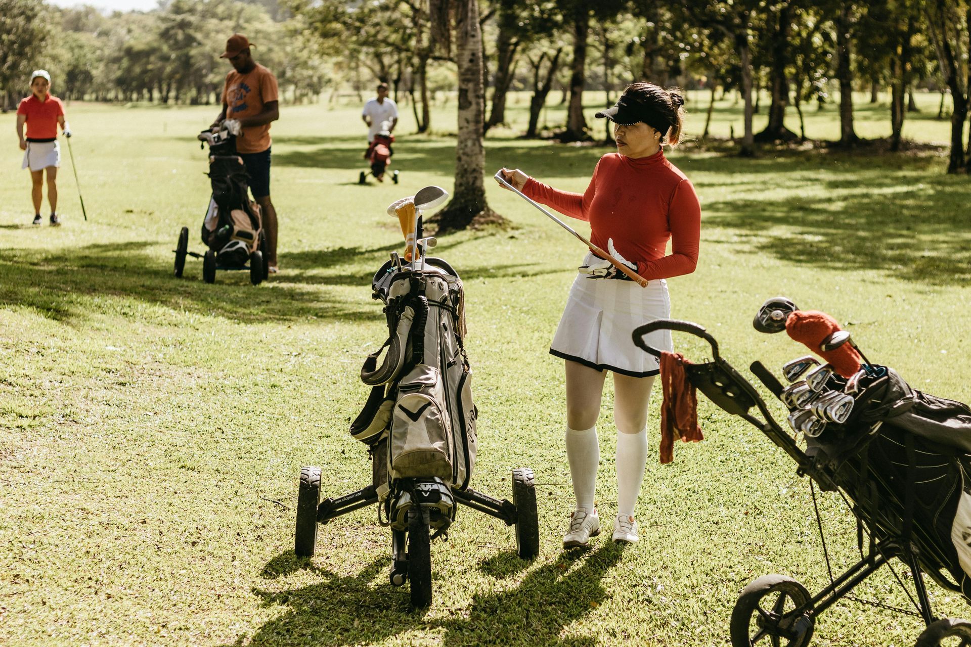 Woman in red golf attire adjusts her club near her golf bag. Golfers and golf carts are visible on a green fairway.