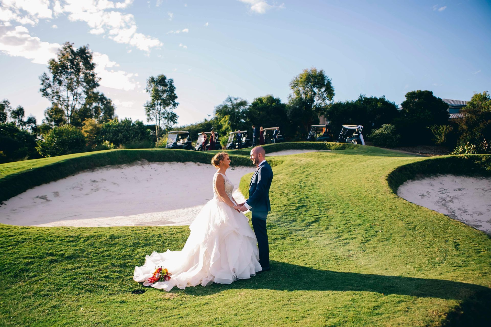 Bride and groom holding hands on a golf course; the bride in a white gown, the groom in a dark suit. They stand between two sand traps.