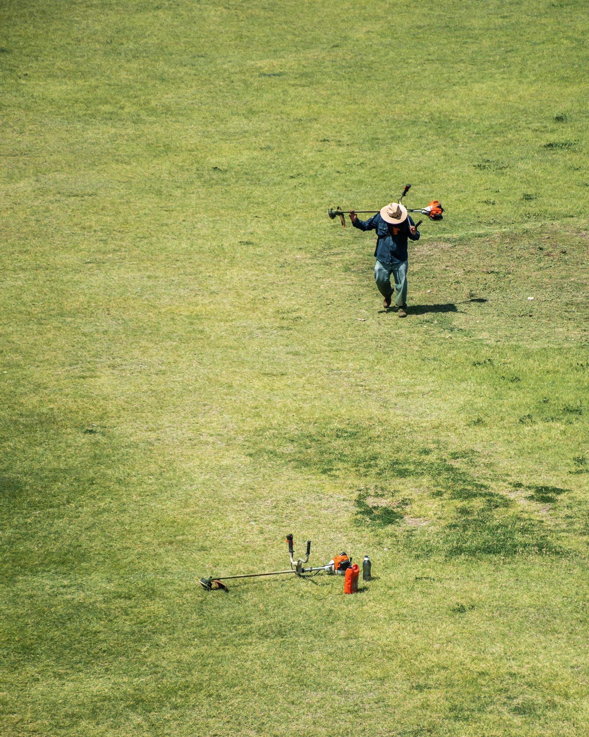 Man with tools on his shoulder walks across a grassy field.