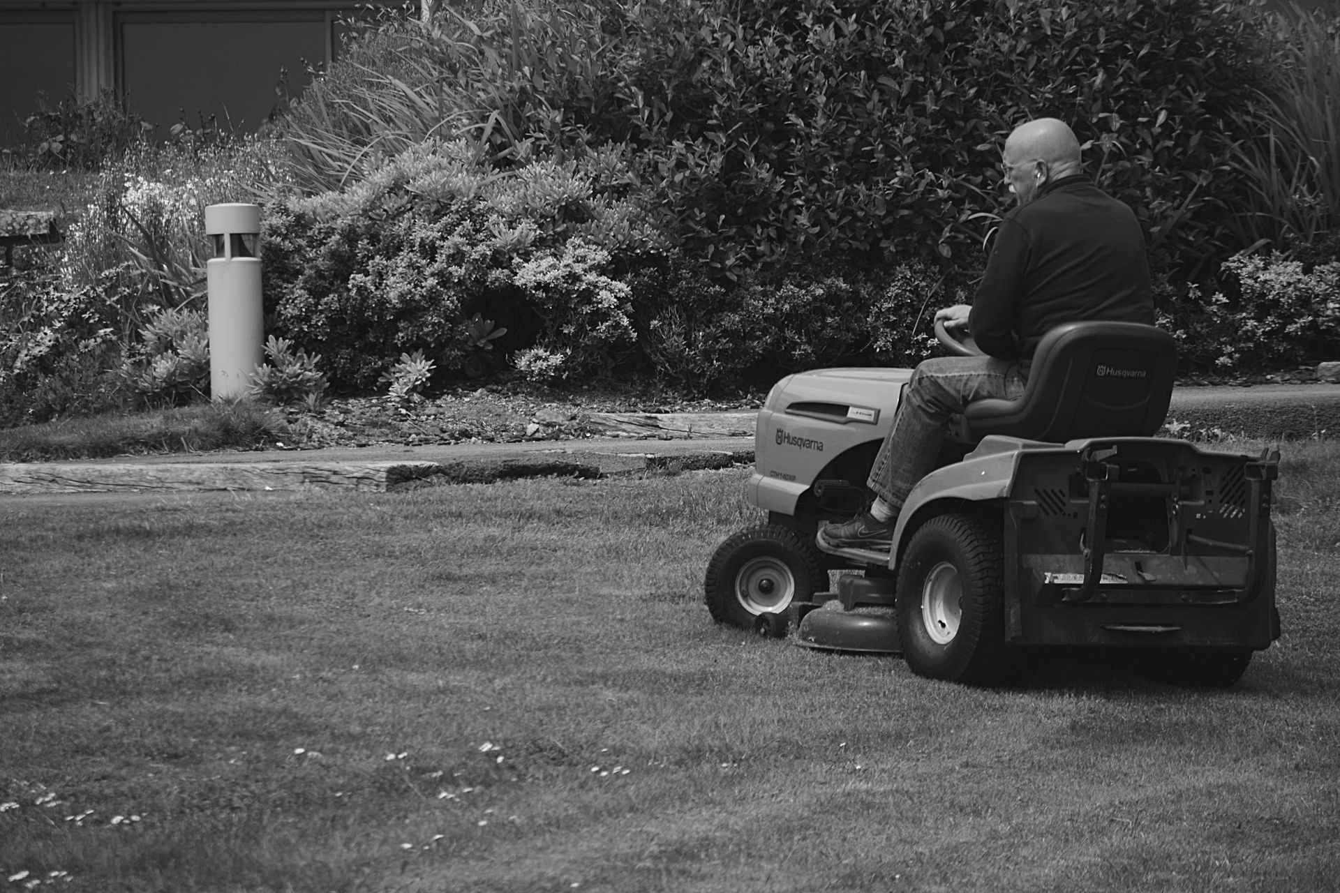 Man on a riding lawnmower mowing grass in front of a building and bushes. Black and white.