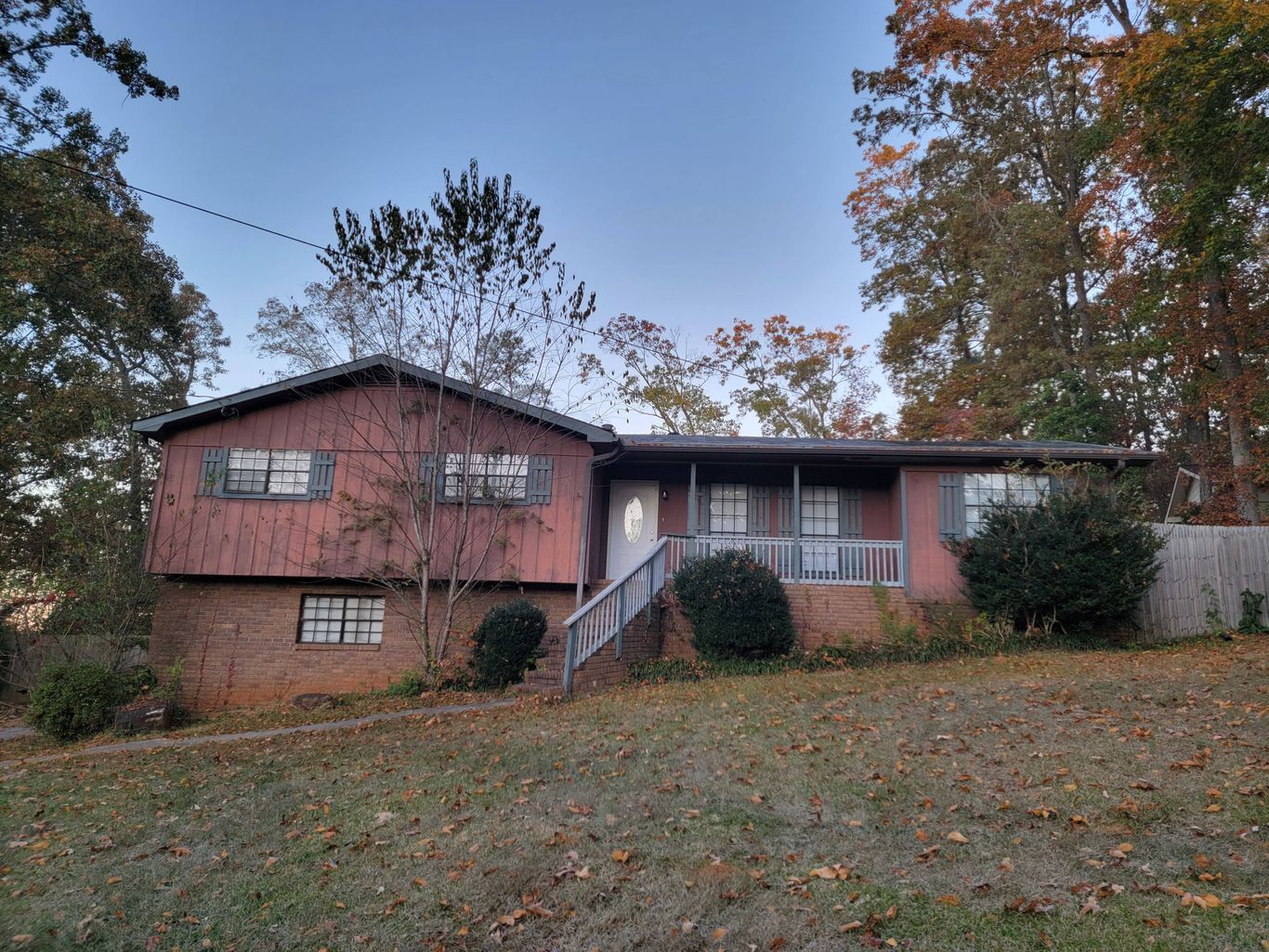 A reddish-brown house with a brick base sits on a grassy hill; fall foliage surrounds it under a blue sky.
