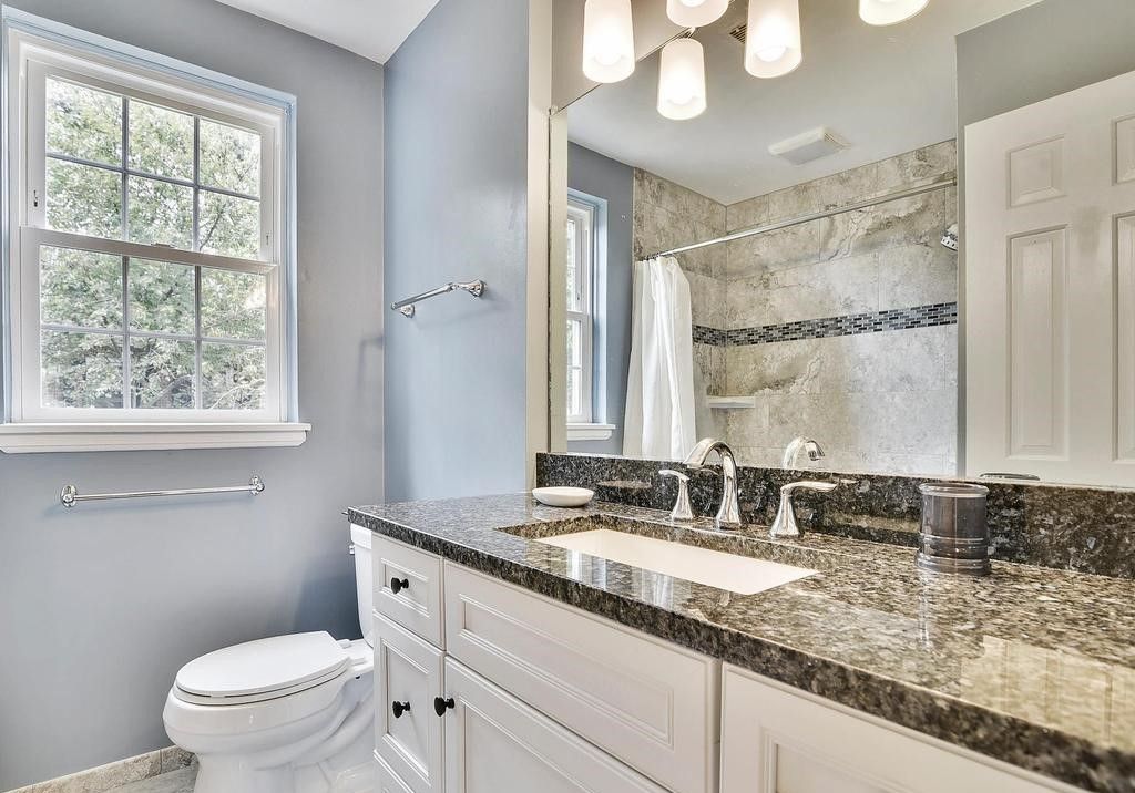 A bathroom with a toilet , sink , mirror and window.