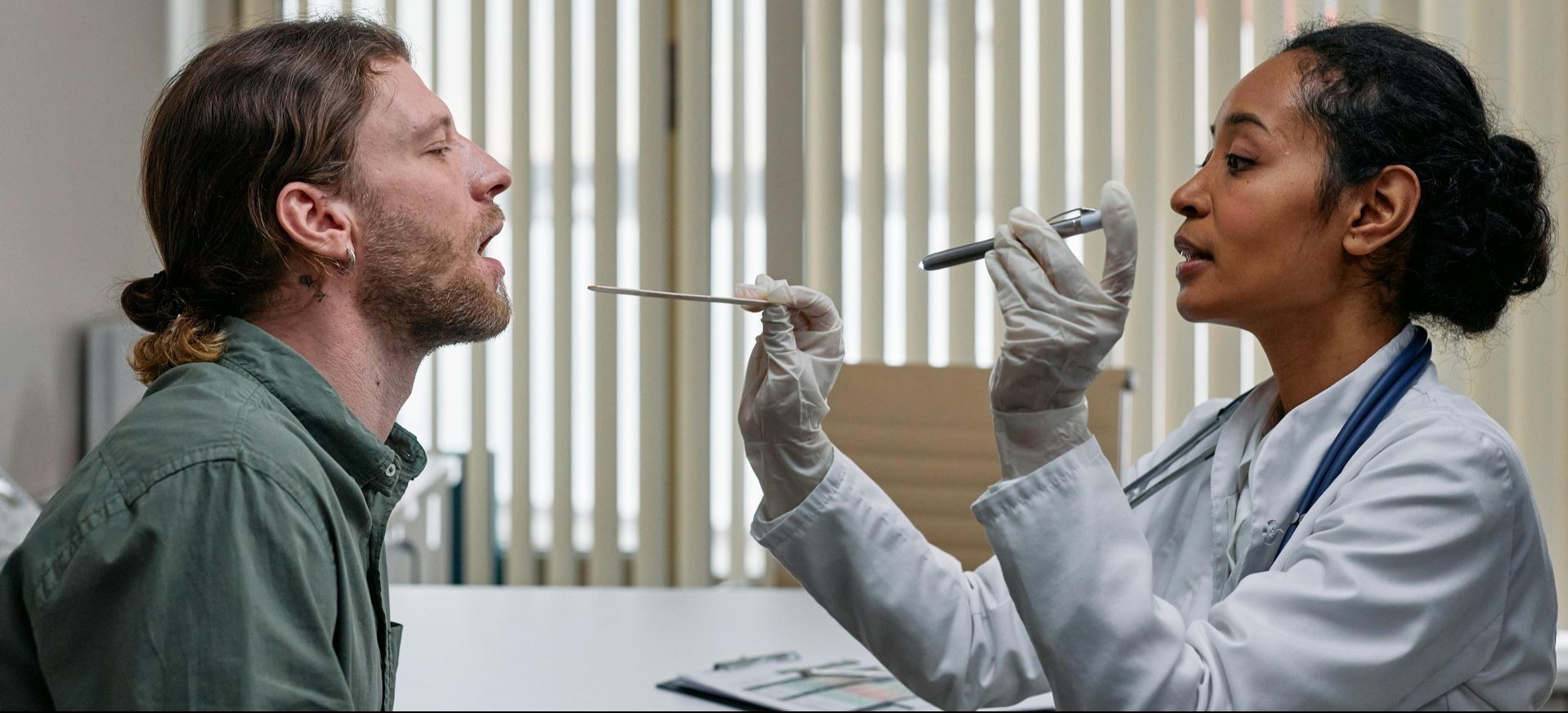 person in white medical jacket holds a medical flashlight and looks in the mouth of a patient