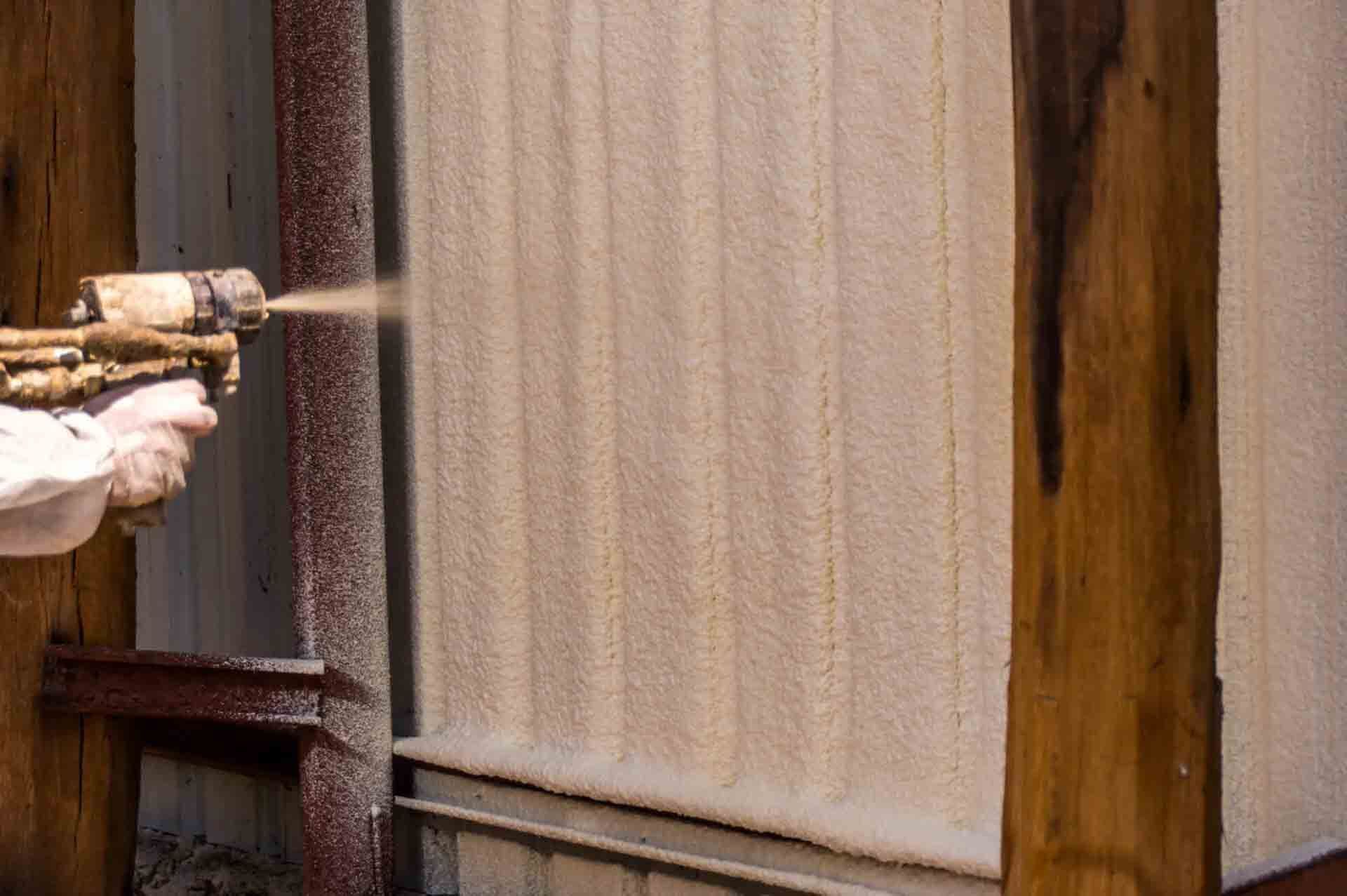 Sprayed insulation being applied to a wall by a person using a spray gun. Wooden beams are visible.