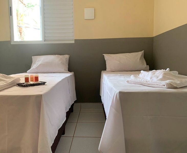 Um quarto com duas camas de solteiro, lençóis brancos, travesseiros e toalhas dobradas, em uma parede bicolor cinza e creme.