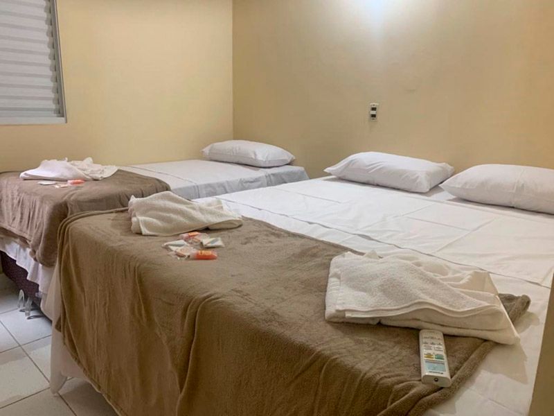 Duas camas com lençóis brancos e cobertores bege em um quarto simples com paredes bege-claras e uma janela com persianas.