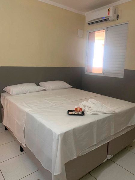 Um quarto limpo com cama de casal com lençóis brancos, uma mesa de cabeceira, janela com persianas brancas e ar condicionado.