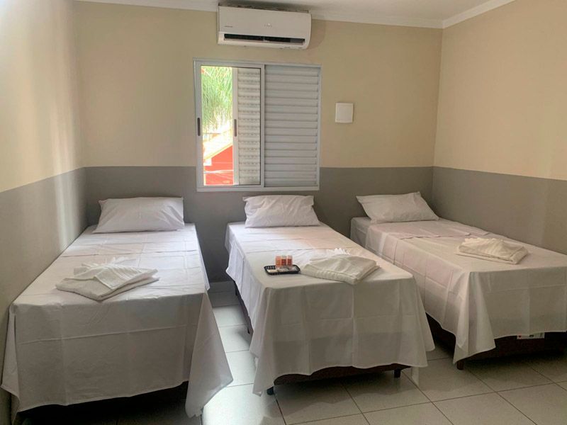 Um quarto de hotel com três camas de solteiro, lençóis brancos, paredes claras e uma janela com persianas sob um ar-condicionado.