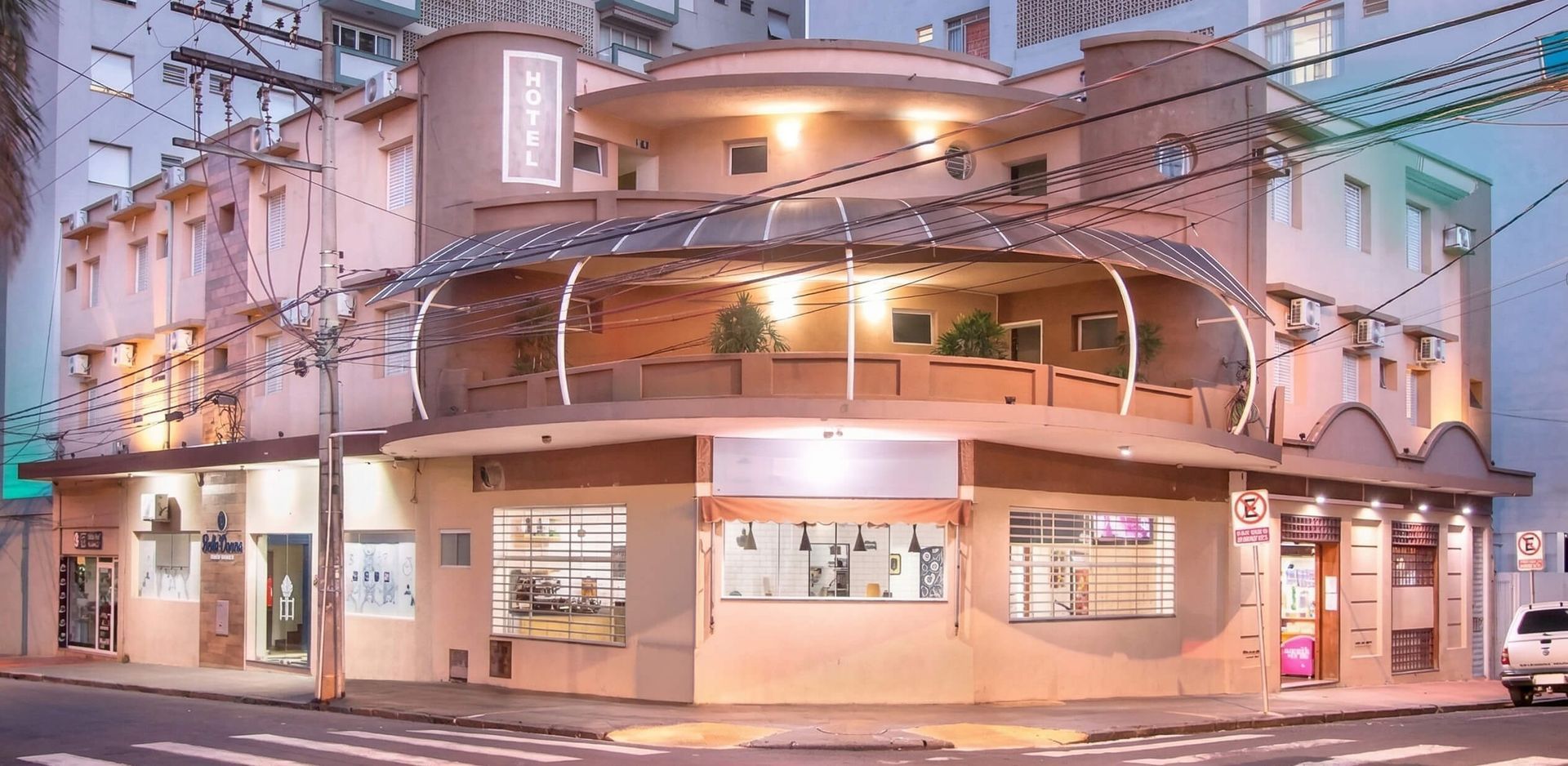 Vista da esquina, ao nível da rua, de um prédio bege com fachada iluminada e varanda em arco durante o crepúsculo.