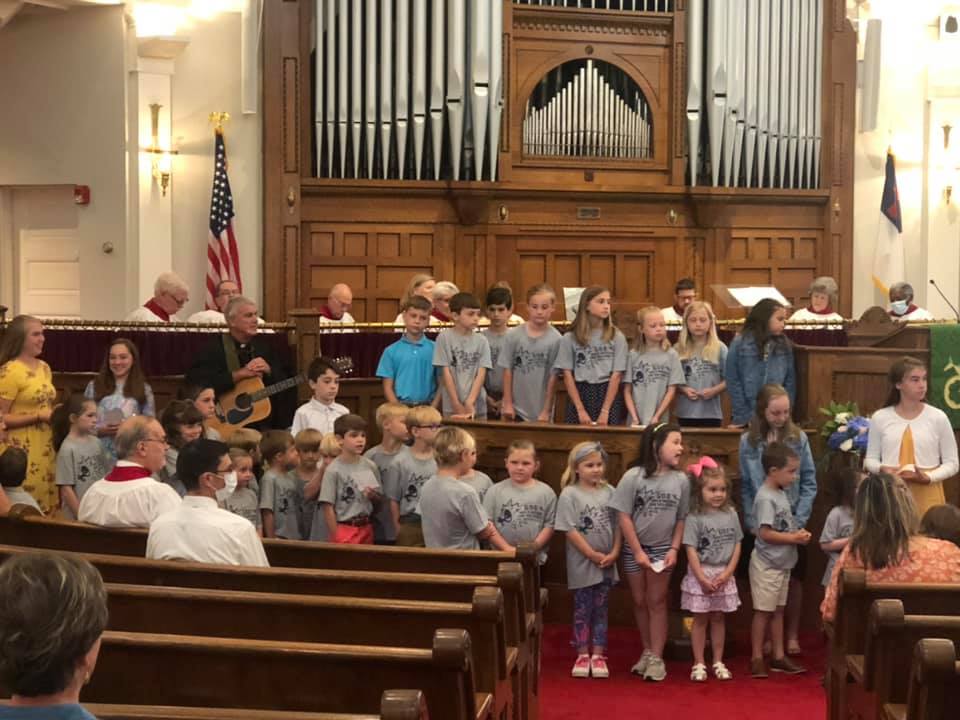 Children's choir singing in a church, organ pipes behind them, with adults observing and playing music.