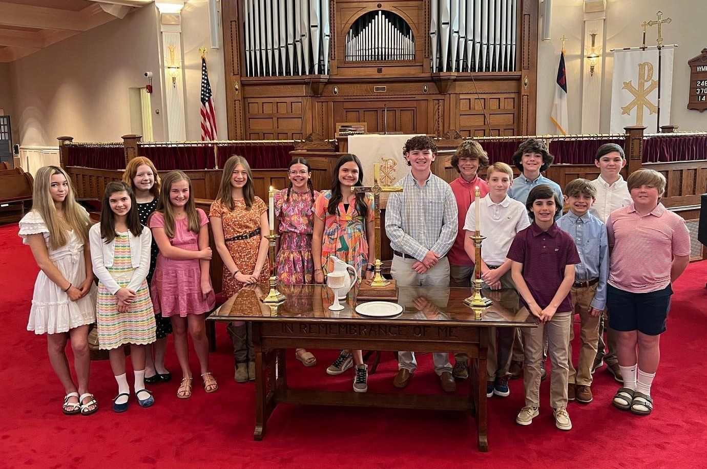 Group of teens pose behind a table in a church, facing the camera. Red carpet and wooden organ backdrop.