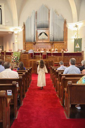 A child in a white robe walks down a red carpet in a church. Pews, organ, and people are visible.