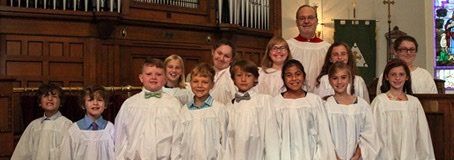 Children in the church's Covenant Choir  in white robes with the Director of Music in a red collar stand in a church.