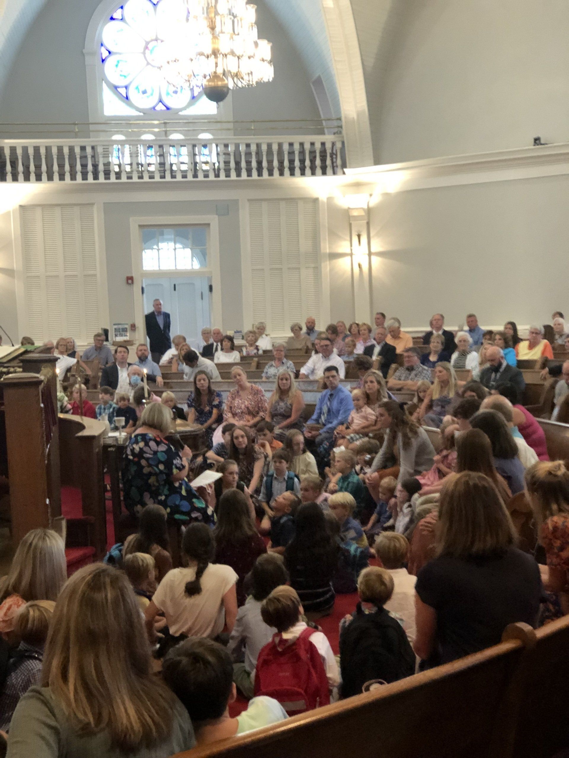 People gathered in a church, some seated. A woman speaks in front, with a balcony overhead.
