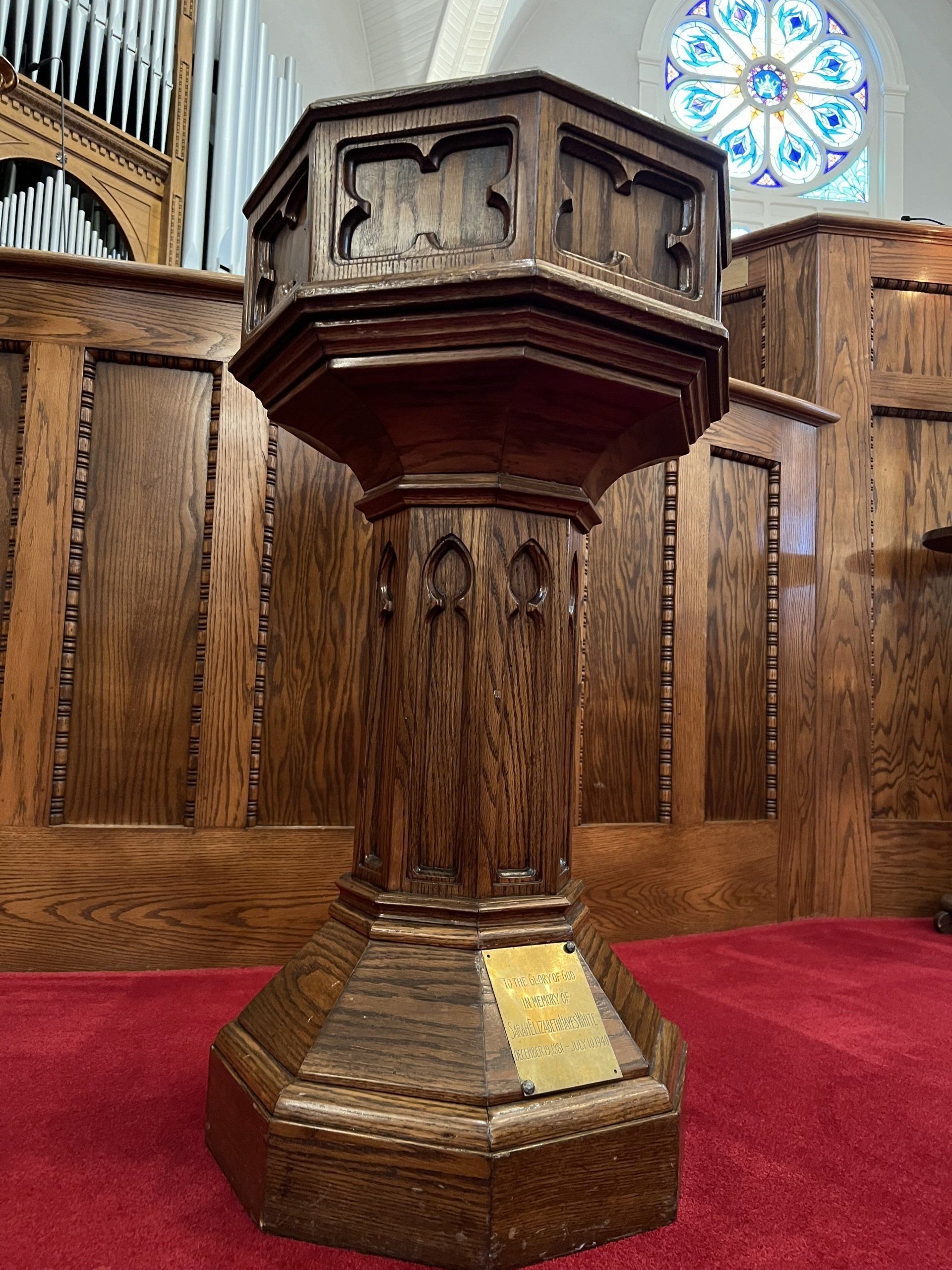 Wooden baptismal font in the Sanctuary of FPC Rock Hill, octagonal shape, ornate carvings,  stained glass in background.